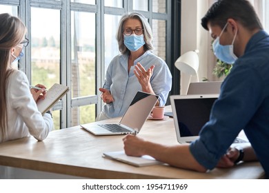 Senior older educator working with computers in office with team wearing medical masks during covid lockdown.Adult experienced business woman is holding meeting with female assistant and male manager. - Powered by Shutterstock