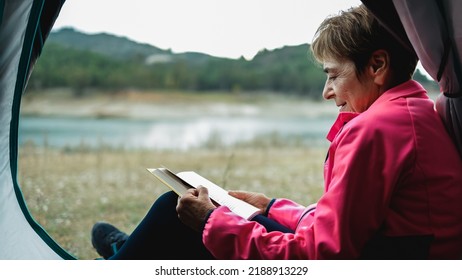 Senior Older Woman Camping While Having Leisure Time Reading A Book In Front Of Lake Mountain - Travel Concept