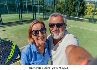 senior padel couple, both wearing sunglasses, celebrates their victory by taking a selfie on the court. Smiling and proud, they capture the joyful moment right after winning their padel game - Powered by Shutterstock