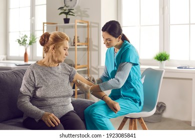 Senior Patient Getting Vaccine Shot Sitting On Sofa At Home. Young Female Nurse Or Doctor Giving Arm Injection To Mature Woman Who's Sitting On Couch. Vaccination And Immunization For Old People