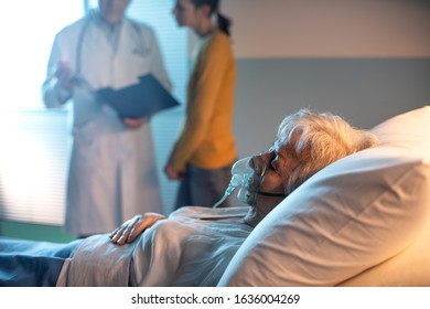 Senior Patient At The Hospital Lying In Bed And Doctor Talking With A Family Member In The Background