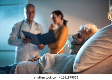 Senior Patient At The Hospital Lying In Bed And Doctor Talking With A Family Member In The Background