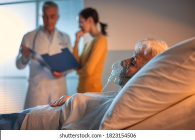 Senior Patient At The Hospital Lying In Bed And Doctor Talking With A Family Member In The Background