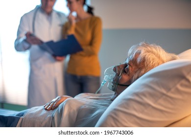 Senior Patient At The Hospital Lying In Bed And Doctor Talking With A Family Member In The Background