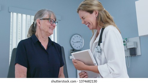 Senior Patient Laughing With Her Doctor In Exam Room. Female Primary Care Physician Talking To Older Caucasian Woman During Regular Check Up