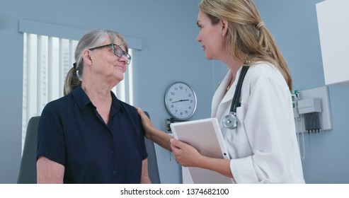 Senior Patient Laughing With Her Friendly Reassuring Doctor In Exam Room. Female Primary Care Physician Talking To Older Caucasian Woman During Regular Check Up