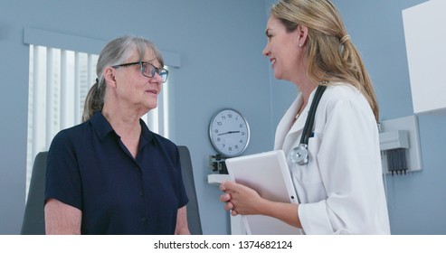 Senior Patient Laughing With Her Friendly Reassuring Doctor In Exam Room. Female Primary Care Physician Talking To Older Caucasian Woman During Regular Check Up