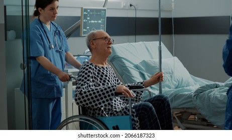 Senior Patient Leaving Hospital Ward In Wheelchair For Surgery Preparations. Nurse And Doctor Helping Old Person With Oximeter And IV Drip Bag Going To Emergency Room With Modern Equipment