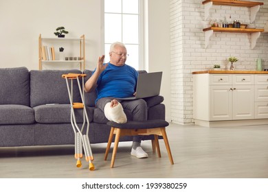 Senior Patient With Physical Injury Having Video Call With Family Or Consultation With Online Doctor. Adult Man With Broken Leg In Cast Sitting On Sofa With Crutches And Waving Hand At Laptop Computer