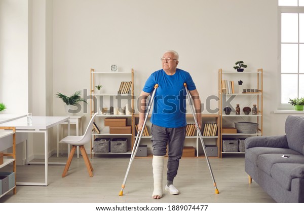 Senior patient successfully undergoes\
rehabilitation after injury in home or car accident: Old man with\
broken leg in plaster cast making good progress and walking in the\
living-room, using\
crutches