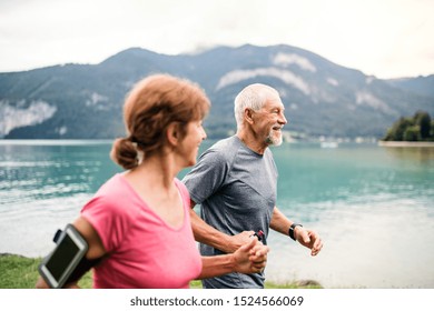 Senior Pensioner Couple With Smartphone Running By Lake In Nature.