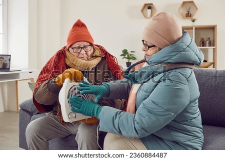 Senior pensioner family couple warming up by electric heater at home. Mature man and woman in winter coat jackets sitting together on sofa couch with small fan heater in their very cold flat or house