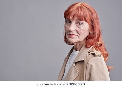 Senior People. Fashion And Beauty. Portrait Of A Good-looking Elderly Woman With Bright Red Hair Dressed In Modern Clothes. Studio Portrait On A Grey Background. Copy Space.