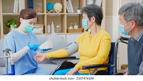 Senior People Home Healthcare Concept - Asian Female Doctor Wearing Face Mask Is Using Blood Pressure Meter On Senior Woman While Elder Man Take Care Of Her