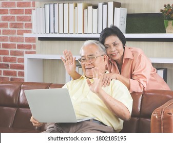 Senior People Lifestyle  Concept. Happy Asian Elderly Couple Making Video Call On Laptop, Senior Man Sitting On Sofa While His Wife Hugging Him From Behind Waving On Computer Screen And Smiling..