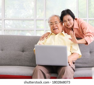 Senior People Lifestyle Concept. Happy Asian Elderly Couple Making Video Call On Laptop, Senior Man Sitting On Sofa While His Wife Hugging Him From Behind Waving On Computer Screen And Smiling.