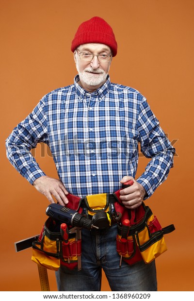 Senior people, occupation and maintenance\
service. Stylish senior retired Caucasian bearded man in his\
sixties posing at orange wall wearing belt with all instruments,\
earning money, doing\
renovations
