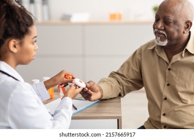 Senior People Support Concept. Portrait Of Black Medical Worker Holding Personal Alarm Button And Giving It To Mature Patient, Sitting At Table In Office, Explaining How To Use Medical Alert