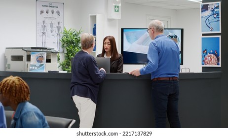 Senior people talking to receptionist at hospital reception desk, asking about medical appointment and healthcare system. Old patients sitting at reception counter in waiting room lobby. - Powered by Shutterstock