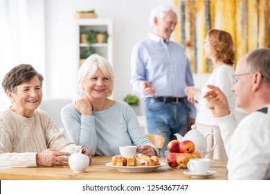 Senior People Visiting Old Friends At Home Sitting Together At Wooden Table Chatting, Drinking Tea And Eating Cake