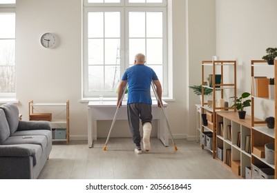 Senior Person With Injured Foot Using Crutches And Walking On His Own At Home. Back View Of Lonely Elderly Man With Broken Leg In Gypsum Plaster Cast Standing In Living Room And Looking Through Window