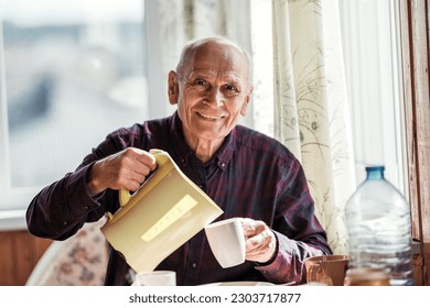 Senior person pouring hot water in cup and smiling seated at kitchen table indoor of rustic house - Powered by Shutterstock