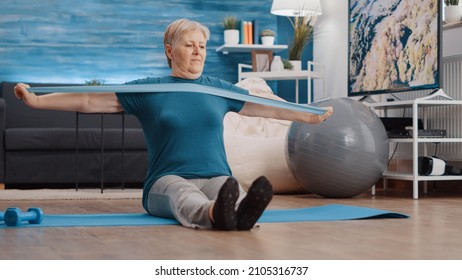 Senior Person Pulling Resistance Band To Exercise On Yoga Mat. Aged Woman Using Elastic Belt To Do Workout Training At Home, Stretching Arms Muscles. Old Adult Exercising With Equipment