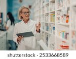 Senior pharmacist wearing uniform and holding medicine bottle and using digital tablet for checking prescription in pharmacy