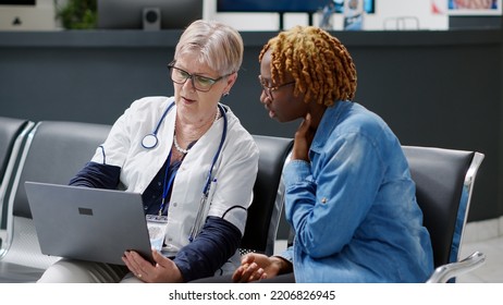 Senior Physician Showing Disease Diagnosis To Young Woman On Laptop, Explaining Treatment At Healthcare Consultation In Medical Center Lobby. Doing Checkup Appointment At Clinic.