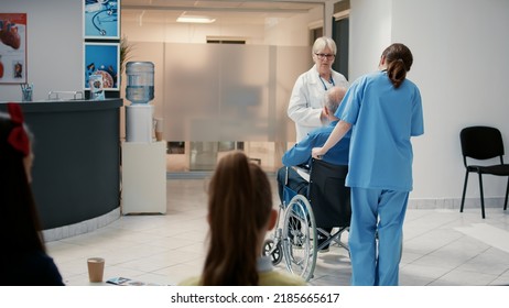 Senior Physician Talking To Retired Man With Disability At Hospital Reception, Preparing To Do Medical Consultation. Wheelchair User In Waiting Room Lobby Attending Checkup Appointment.