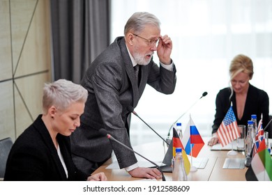 Senior Politician Offers His Plan Of Action And Share Opinion, Elderly Man In Suit Talking To Multi-ethnic Group Of Partners Sitting At Desk In Boardroom, Discussing