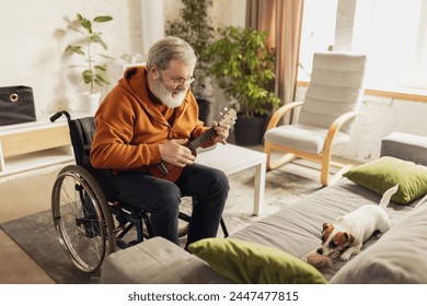 Senior positive man sitting on wheelchair at home in cozy warm living room and playing ukulele guitar in front of little dog playing on sofa. Concept of healthcare, lifestyle, wellness, empowerment - Powered by Shutterstock