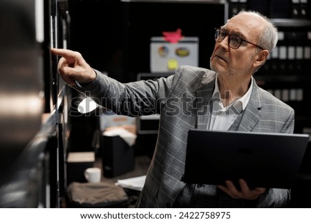 Senior private detective in criminal cases archive room looking for criminal case file details on cabinet shelves. Elderly investigator surrounded by criminology folders in agency depository office