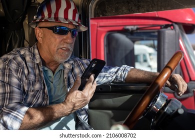 Senior Professional Truck Driver Wearing Hard Hat And Sunglasses Sits In The Cabin And Talks On The Smartphone. Elderly Trucker Inside Big Rig At The Steering Wheel Talking To Dispatcher On Cell Phone