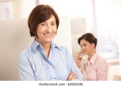 Senior Professional Woman Smiling At Camera, Assistant Using Land Line Phone In Background.