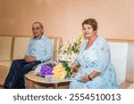 A senior Russian couple, 71 and 75 years old, just married at the registry office. They sit beside floral bouquets, both in blue attire, symbolizing family values and love