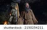 Senior sage man turning around in cave. Elderly bearded male hermit in hooded brown robe with staff turning around and looking at camera near stone walls with candles in cave