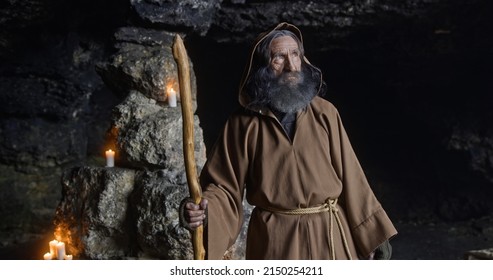 Senior sage man turning around in cave. Elderly bearded male hermit in hooded brown robe with staff turning around and looking at camera near stone walls with candles in cave - Shutterstock ID 2150254211