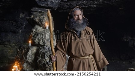Senior sage man turning around in cave. Elderly bearded male hermit in hooded brown robe with staff turning around and looking at camera near stone walls with candles in cave