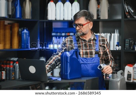 Senior salesman using barcode scanner and laptop, verifying motor oil product information in auto parts store, surrounded by shelves full of car maintenance products