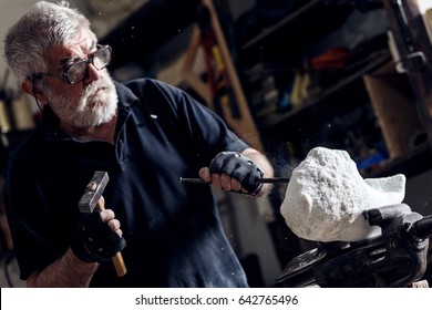 Senior sculptor working on his marble sculpture in his workshop with hammer and chisel.