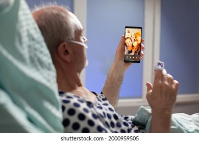 Senior Sick Man Breathing Through Oxygen Tube Saying Hello To Relatives, Laying In Hospital Bed Holding Phones, Discussing About Recovery. Old Man Waiting For Treatment.