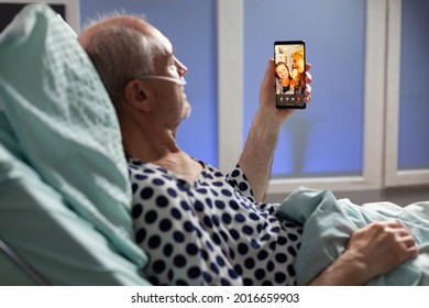 Senior Sick Man Breathing Through Oxygen Tube Saying Hello To Relatives, Laying In Hospital Bed Holding Phones, Discussing About Recovery. Old Man Waiting For Treatment.