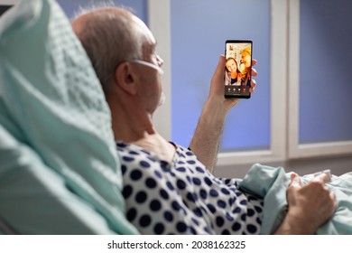 Senior Sick Man Breathing Through Oxygen Tube Saying Hello To Relatives, Laying In Hospital Bed Holding Phones, Discussing About Recovery. Old Man Waiting For Treatment.