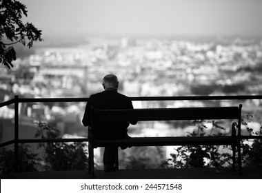 Senior is sitting alone on the bench looking at city at evening. A lonely old man sitting on a bench in a park.Elderly gentleman on park bench in contemplation,black and white photo