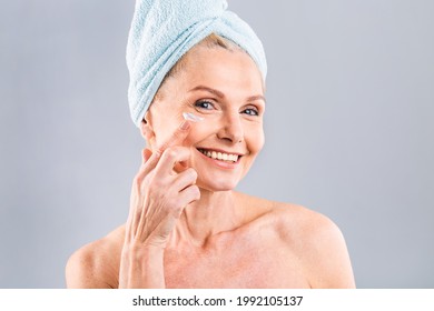 Senior Smiling 50s Middle Aged Mature Older Woman Applying Facial Cream On Face Looking At Camera Isolated Over White Background. Anti Age Healthy Dry Skin Care Beauty Therapy Concept.