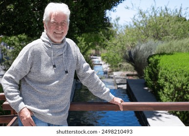 Senior smiling bearded man  in a cozy sweater looks at camera enjoying a peaceful sunny day outdoors, green park background - Powered by Shutterstock