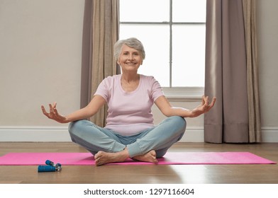 Senior smiling woman doing yoga in her living room. Elderly relaxed woman sitting in lotus pose and meditating while practicing yoga at home. Old grandmother sitting on yoga mat smiling.