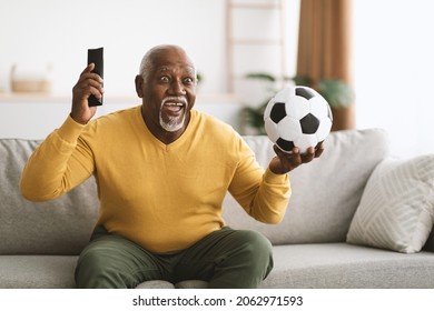 Senior Soccer Fan. Mature Black Man Watching Sport Channel On TV Holding Football Ball And Remote Control, Shouting Celebrating Victory And Cheering Team Sitting On Sofa At Home