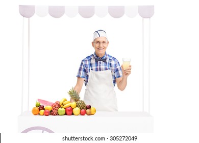 Senior Soda Jerk Holding A Glass Of Lemonade And Standing Behind A Stall With A Bunch Of Fruits On It Isolated On White Background